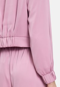 Lichtroze blouse met een verzamelde taille, lange puffmouwen en een soepele, zijdeachtige textuur. De stof heeft subtiele verticale plooien.