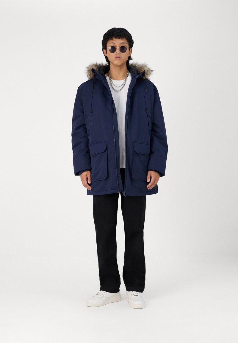 Marine Parka mit Fellbesatz an der Kapuze, Frontreißverschluss und zwei großen Fronttaschen, kombiniert mit schwarzen Hosen und weißen Turnschuhen.