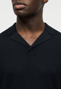 Κοντινό πλάνο ενός ατόμου που φοράει μαύρο πλεκτό πόλο μπλουζάκι με υφή, ανοιχτό γιακά και ένα μόνο ορατό κουμπί.