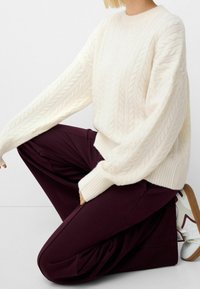 Chunky cable-knit sweater in cream, with ribbed cuffs and hem, worn with dark burgundy trousers. Model kneeling, paired with white sneakers.