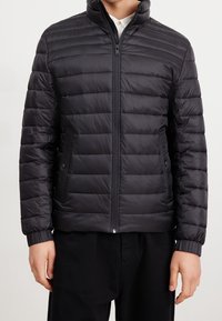 Muž oblečený v černé prošívané bundě na zip s bočními kapsami a elastickými manžetami přes bílé tričko.