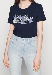 Námořnické modré tričko s květinovým potiskem a nápisem "Lauren Ralph Lauren". Krátké rukávy, kulatý výstřih, měkký bavlněný materiál. Spárováno se světle modrými džíny.