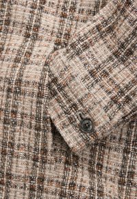 Gros plan d'un tissu à carreaux marron, beige et blanc avec un tissage texturé et un bouton rond foncé sur le poignet d'une manche de veste.
