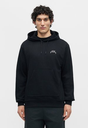 Άνδρας με κατσαρά μαλλιά που φοράει μαύρο φούτερ με κουκούλα και το λογότυπο ACW στο αριστερό μέρος του στήθους και μαύρο παντελόνι, στέκεται μπροστά σε απλό ανοιχτό φόντο.