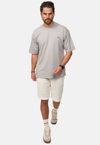 Helles graues Baumwoll-T-Shirt mit kurzen Ärmeln und kleinem bunten Logo auf der Brust, kombiniert mit weißen strukturierten Shorts und beigen Sneakern.