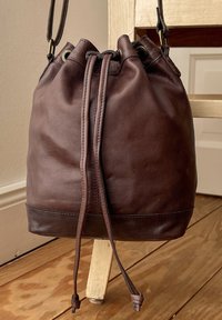 Borsa a secchiello in pelle marrone con chiusura a coulisse, texture liscia e base in pelle più scura. Presenta occhielli in metallo e tracolle regolabili.