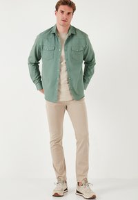 Cămașă verde cu butoni, dotată cu două buzunare pe piept, purtată cu un tricou bej deschis și pantaloni de culoare deschisă, completată de adidași bej.