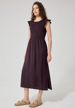 Frau mit lockigem Haar, die ein ärmelloses dunkelviolettes Midikleid und beigefarbene Sandalen mit Schnallen trägt, steht vor einem schlichten hellen Hintergrund.
