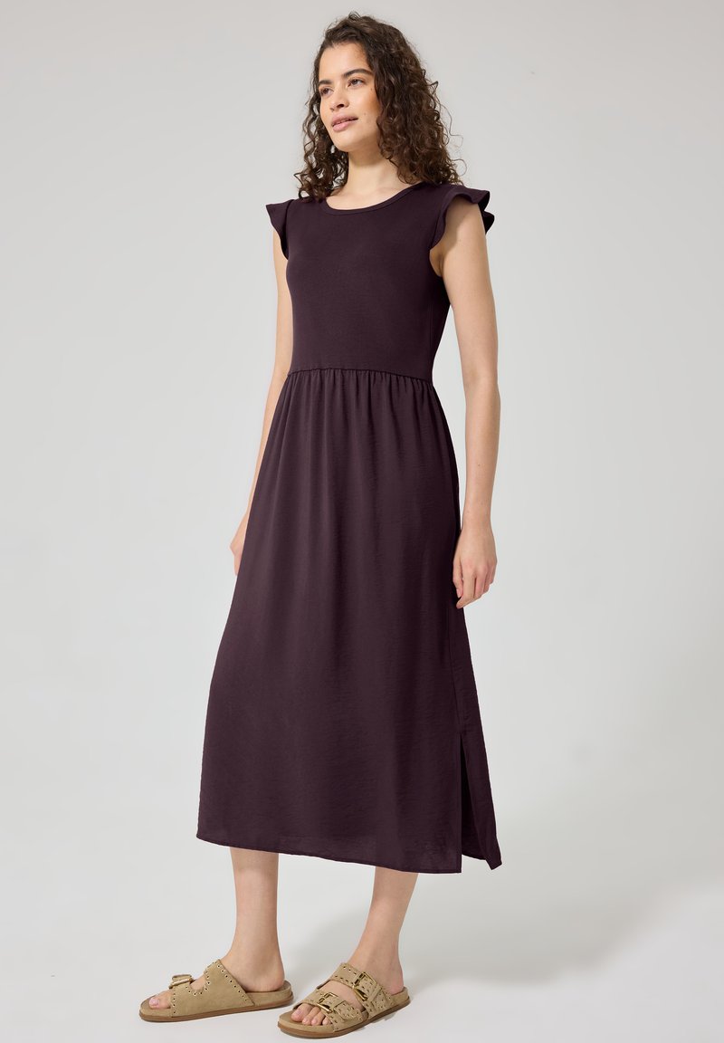 Femme aux cheveux bouclés portant une robe midi sans manches violet foncé et des sandales beige à boucle, debout devant un fond clair uni.