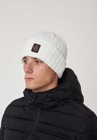 Berretto lavorato a costine bianco con una toppa rettangolare nera che presenta un logo, indossato da una persona in una giacca a vento nera.