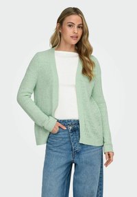 Mint green knitted cardigan with a loose fit, open front, and ribbed cuffs, paired with a white top and high-waisted blue jeans.
