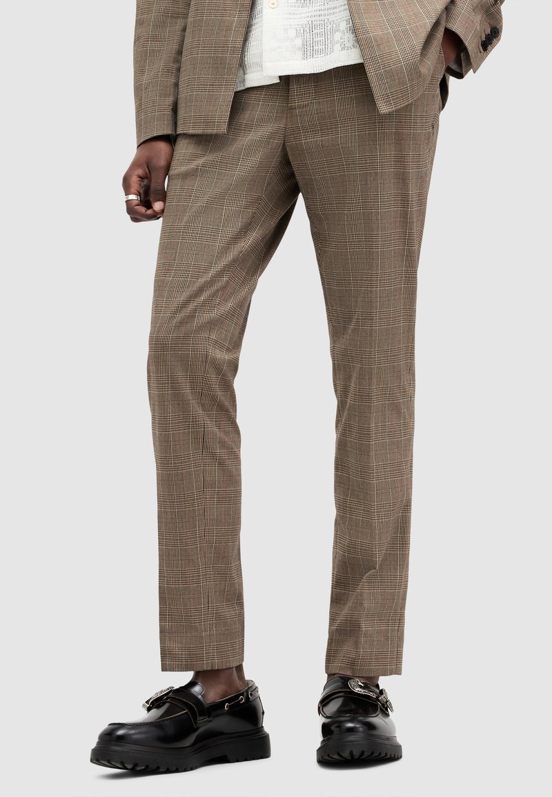 Man wearing brown plaid suit pants and matching blazer, with black leather loafers featuring metal buckle details.