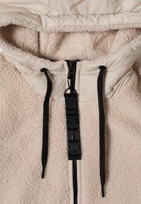 Veste polaire beige à capuche avec cordons noirs et tirette de fermeture éclair noire étiquetée "CECIL" en gros plan.