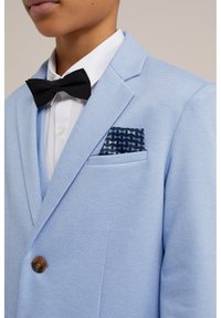 Lichtblauwe blazer met een gestructureerde afwerking, voorzien van een bruine knoop en een marineblauwe pochets met een geometrisch patroon. Witte shirt en zwarte vlinderdas.
