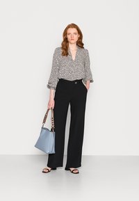Zwarte wijde broek gecombineerd met een patternd blouse met een V-hals en puffmouwen. Een blauwe handtas met een schouderband rust op de heup.