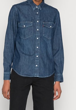 Person iført en mørkeblå denimskjorte med knapper og brystlommer samt sorte bukser, stående mod en ensartet lys baggrund.