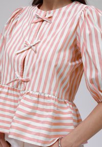 Peplum-Bluse in rosa-weißen senkrechten Streifen, mit Rundhalsausschnitt, kurzen Puffärmeln und drei Schleifenakzenten an der Vorderseite.