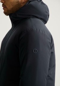 Černá kapucňová bunda s hladkou texturou, která má na rukávu malé vyšívané logo a elegantní design.