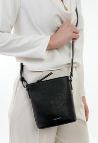 Black leather crossbody bag with a textured finish, rectangular shape, and adjustable strap, held by a person wearing a white top.