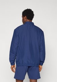 Αντιανεμικό τζάκετ σε ναυτικό μπλε χρώμα με ψηλό γιακά, ελαστικές μανσέτες και στρίφωμα, κατασκευασμένο από ελαφρύ υλικό. Απλός, καθαρός σχεδιασμός, χωρίς εμφανή σχέδια.