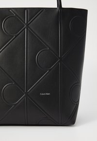 Sac fourre-tout en cuir noir avec des motifs géométriques en relief et un logo. Dispose de longues poignées et d'un design structuré. Texture lisse.
