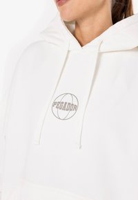 Biely mikina s kapucňou a šnúrkami, s jemným šedým logom. Tkanina má hladký povrch a voľný strih.