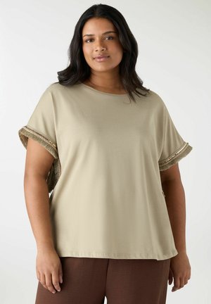 Donna con capelli castani di media lunghezza che indossa una maglia beige a maniche corte con bordi delle maniche con frange e perline e pantaloni marroni.