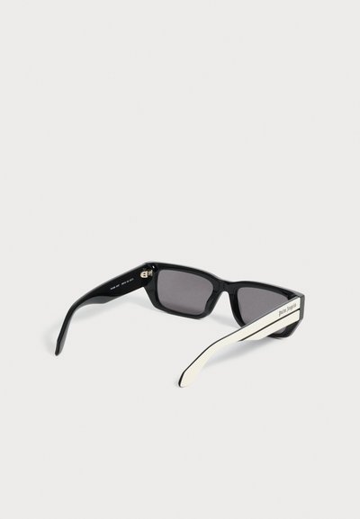 Lunettes de soleil noires rectangulaires avec des verres foncés et des branches blanches comportant un petit texte sur un côté, sur un fond clair uni.