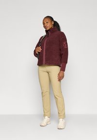 Burgunderfarget fleecejakke med høy krage, glidelås og teksturert stoff; brukt sammen med khaki bukser og beige sneakers.
