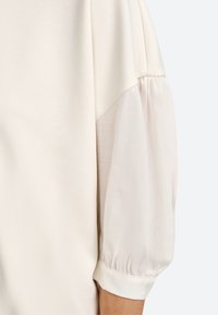 Lys beige top med en blanding af blødt, tekstureret stof og gennemsigtige pufærmer. Har en manchet ved ærmets ende.