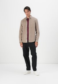 Light brown button-up shirt with a chest pocket, layered over a maroon tee, paired with black pants and white sneakers. Plain background.