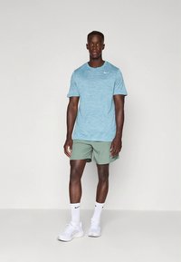 Lys blå atletisk t-skjorte med subtil tekstur og hvit logo, kombinert med grønne shorts, hvite sokker og hvite joggesko.