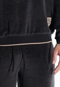 Zwarte velours trainingspak met een licht glanzende textuur, geribbelde boorden en een contrasterende beige rand bij de tailleband, met een label waarop staat "HET GAAT OM HET PLEZIER."