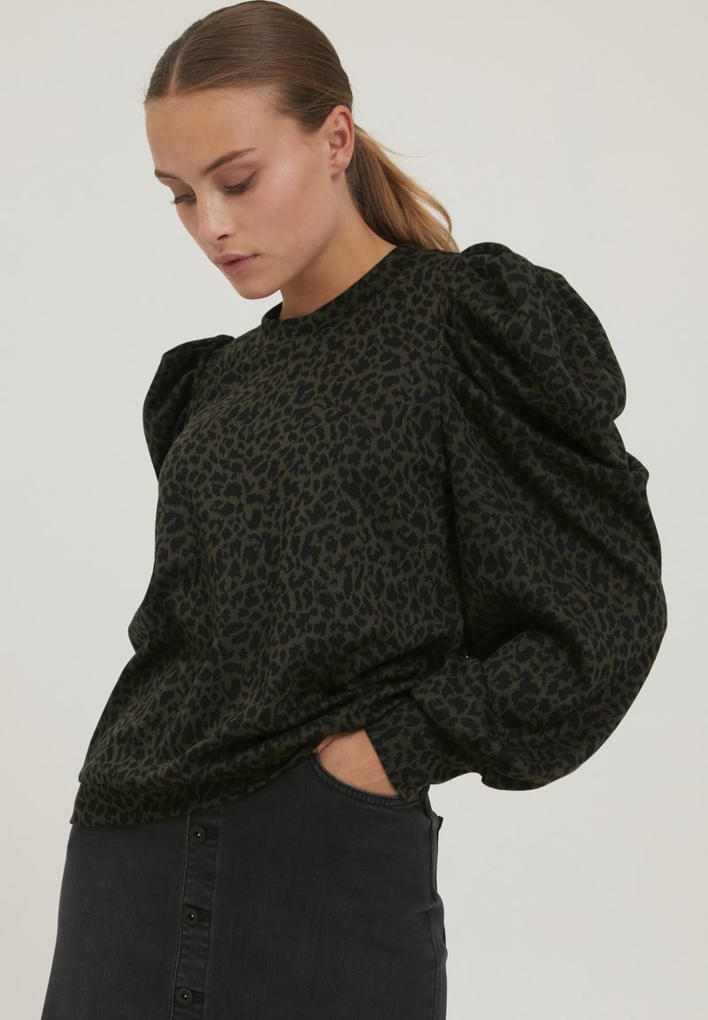 Top negru cu mâneci umflate, având un model subtil de leopard. Textură moale a materialului, guler rotund și croială lejeră. Asortat cu o fustă închisă.