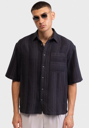 Άνδρας που φορά κοντομάνικο μαύρο πουκάμισο με λευκές κάθετες ρίγες, μπεζ παντελόνι, γυαλιά ηλίου, χρυσό κολιέ και ασημένιο βραχιόλι, ποζάροντας.