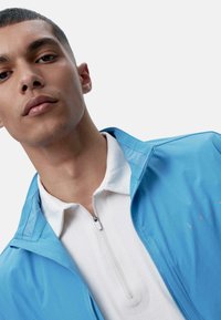 Világoskék, cipzáras dzseki álló gallérral, fehér pólóing felett viselve. Simább textúrájú és minimalista dizájn, finom részletekkel.