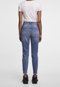 Henkilö, joka käyttää korkeavyötäröisiä sinisiä farkkuja, valkoista lyhythihaista paitaa ja mustia paksupohjaisia urheilukenkiä, seisoo selkä kameraan päin yksinkertaisella taustalla.