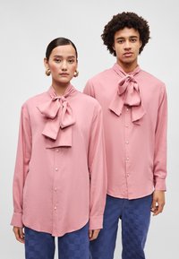 Camisas cor-de-rosa com botões e gravatas borboleta oversized no colarinho. Mangas longas, barra curva, combinadas com calças xadrez azul. Textura suave.