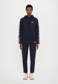 Sudadera navy y joggers a juego hechos de tela suave; presenta detalles del logotipo en blanco a lo largo de las mangas y los pantalones, con un bolsillo tipo canguro.