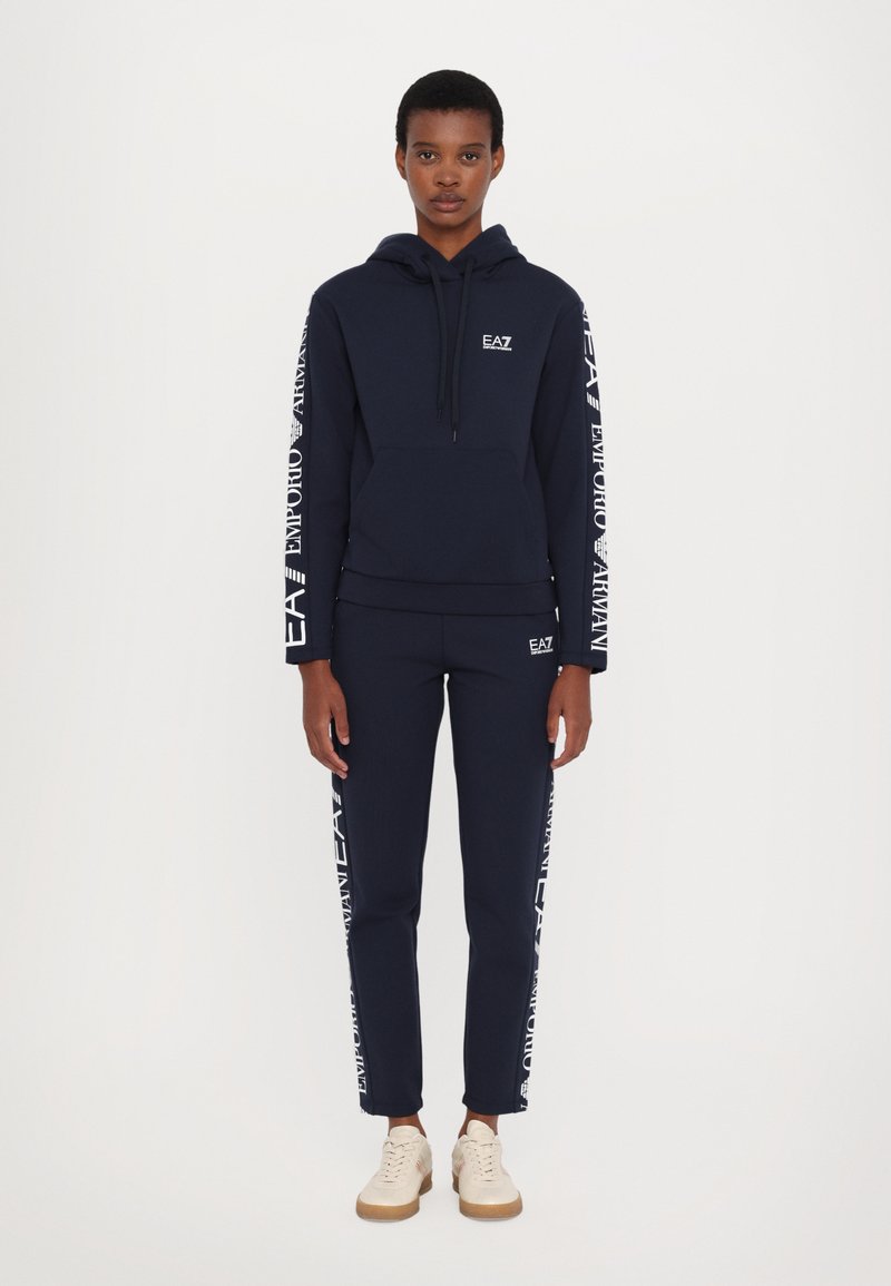 Sudadera navy y joggers a juego hechos de tela suave; presenta detalles del logotipo en blanco a lo largo de las mangas y los pantalones, con un bolsillo tipo canguro.