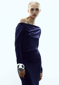 Femme avec les cheveux blonds en arrière, portant une robe bleue marine à épaules dénudées et un grand bracelet réfléchissant en argent à son poignet.