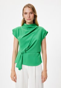 Blusa verde con scollatura drappeggiata e maniche corte, dotata di un dettaglio legato in vita. Tessuto liscio e leggero su uno sfondo semplice.