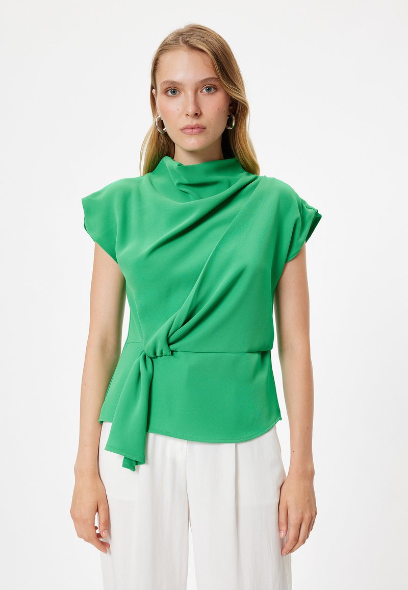 Blusa verde con scollatura drappeggiata e maniche corte, dotata di un dettaglio legato in vita. Tessuto liscio e leggero su uno sfondo semplice.