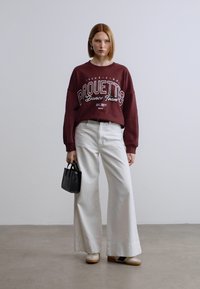 Μακρυμάνικη φούτερ σε μπορντό με λευκά γράμματα; λευκά παντελόνια με φαρδιά μπατζάκια; μικρή μαύρη τσάντα χειρός; παπούτσια σε ανοιχτή καφέ απόχρωση; απαλές υφές και casual στυλ.