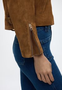 Μανίκι καφέ σουέντ μπουφάν με χρυσό φερμουάρ μερικώς ανοικτό δίπλα σε χέρι που ακουμπά στην τσέπη μπλε τζιν.