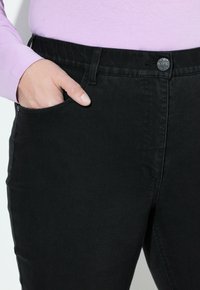 Los pantalones de mezclilla negros cuentan con una textura suave, una silueta ajustada y un bolsillo delantero. La cinturilla incluye un botón de metal y lazos para el cinturón.