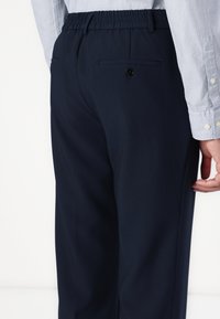 Dunkelblaue Hose mit elastischem Bund, ausgestattet mit einer Gesäßtasche mit Knopf und einer glatten Textur. Sichtbare Hand, die auf der Hüfte ruht.