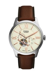 Cinturino in pelle marrone, cassa in acciaio inossidabile argentato, quadrante crema con dettagli in oro rosa, movimento visibile e indicatori in numeri romani.