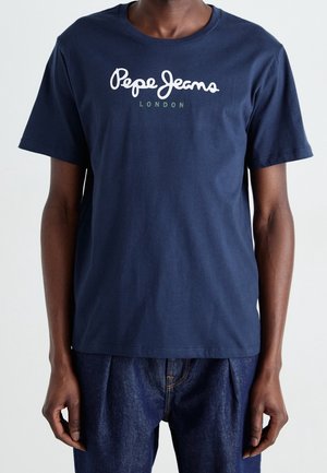 Mann trägt ein marineblaues Pepe Jeans London T-Shirt mit kurzen Ärmeln und dunkelblaue Jeans, Hände seitlich ruhend, vor einem schlichten hellen Hintergrund.