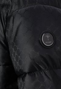 Chaqueta acolchada negra con un tejido texturizado y un diseño patrón sutil. Incluye un emblema redondo negro etiquetado "JOOP!" en la manga.